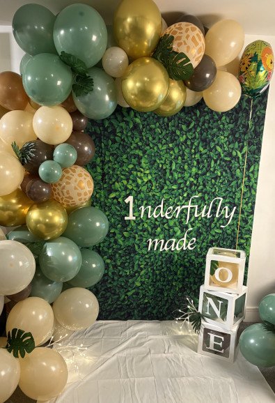 A lush green hedge backdrop featuring a white "1nderfully made" sign, framed by an organic balloon garland in sage green, gold, and giraffe print for a first birthday.