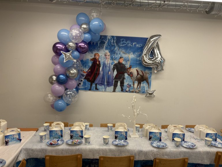 Frozen-themed party setup featuring a character backdrop and  balloon garland. Children’s party table with a froze tree centerpiece, personalised Frozen-themed party boxes 
And paper plates on a snow-effect runner.
