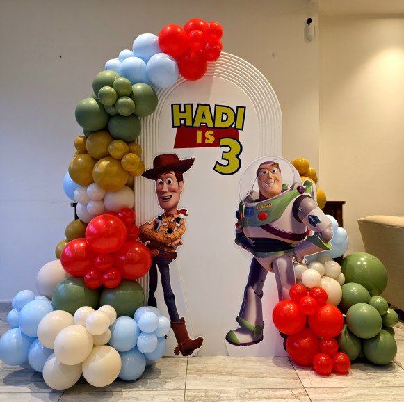 Toy Story birthday backdrop with Woody and Buzz Lightyear cutouts and a red, blue, green and yellow balloon garland framing a white arch.