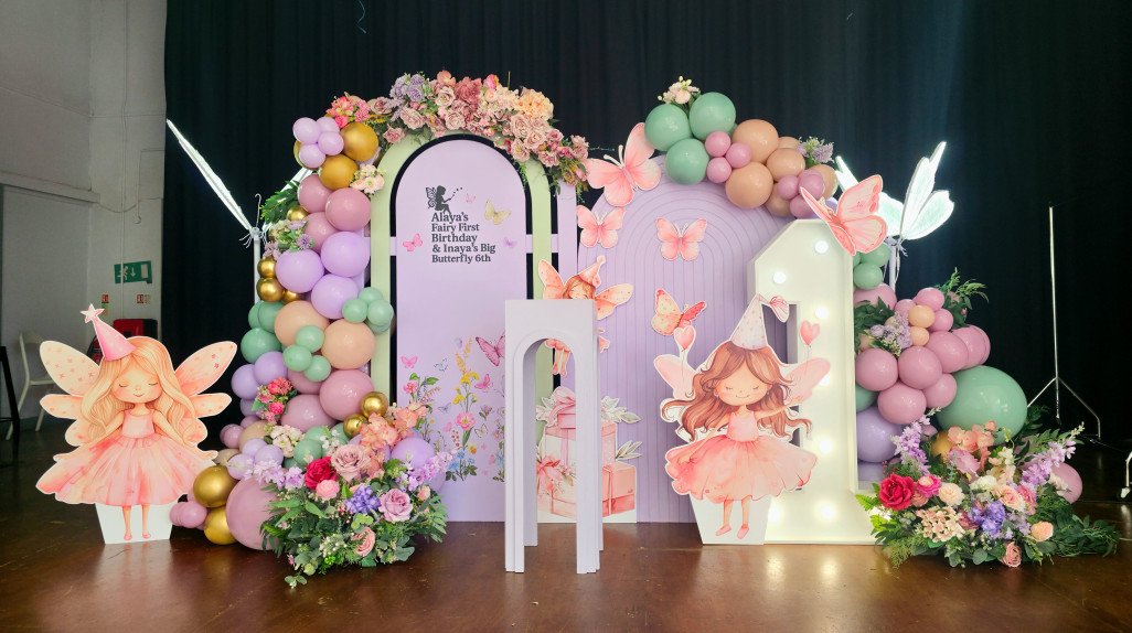 Full fairy and butterfly themed stage setup with pastel decor, cut-outs, flowers and signage celebrating Alaya turning 1 and Inaya turning 6.