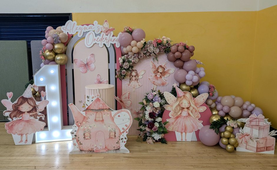 Fairy-themed 1st birthday setup with butterfly backdrop, pink and gold balloon garland, floral accents, and fairy cut-out holding heart balloons.