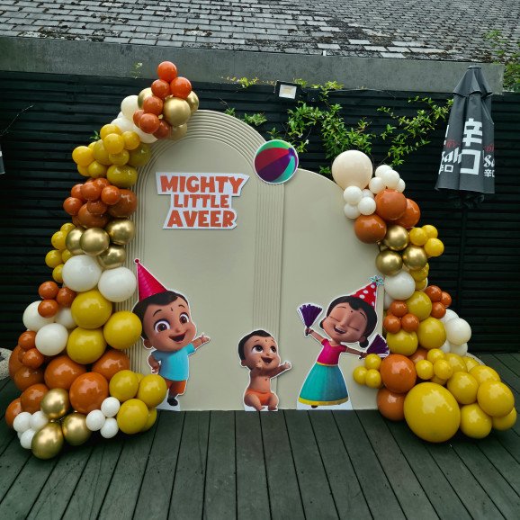 Front view of Little Beam birthday styling at Chin Chin Pinner with playful cartoon cutouts, party hats, and balloon garlands in orange, gold, white, and yellow.