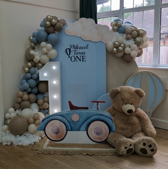 Front view of teddy bear and hot air balloon themed first birthday decor with pastel balloons, number 1 light prop, and blue car prop.