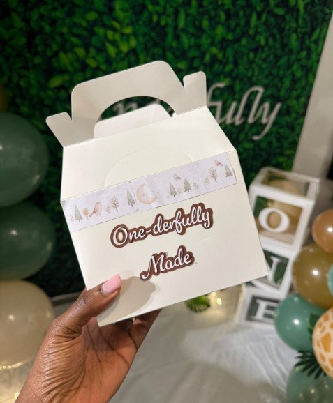 A white party favour box decorated with woodland themed washi tape and a sticker reading "One-derfully Made" for a first birthday celebration.