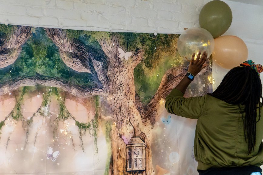 An event stylist reaching up to arrange green and gold balloons against a large enchanted tree tapestry backdrop with twinkling fairy lights.